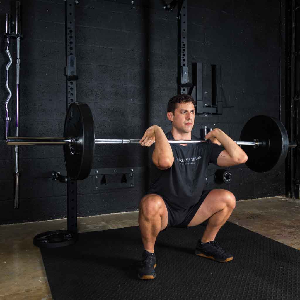 Man performing front squat with Barbell Standard Olympic barbell loaded with plates, bar resting across shoulders in gym, wearing black Barbell Standard shirt and black shoes