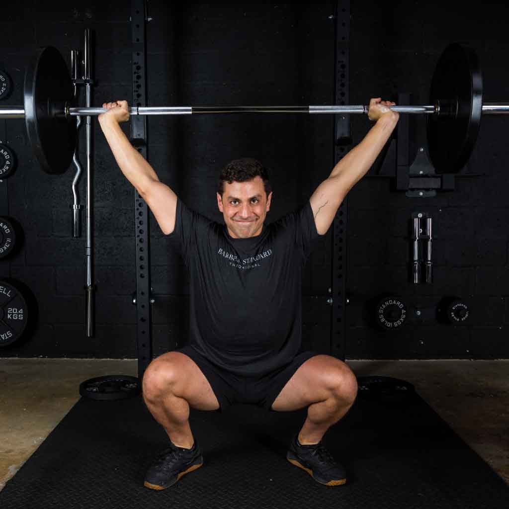 Man performing overhead squat with Barbell Standard Olympic barbell loaded with plates in gym, wearing black Barbell Standard shirt and black shoes.