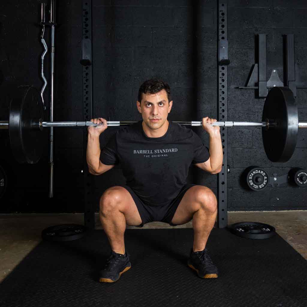 Man performing deep squat with Barbell Standard Olympic barbell loaded with plates in a gym, wearing black Barbell Standard shirt and black shoes.