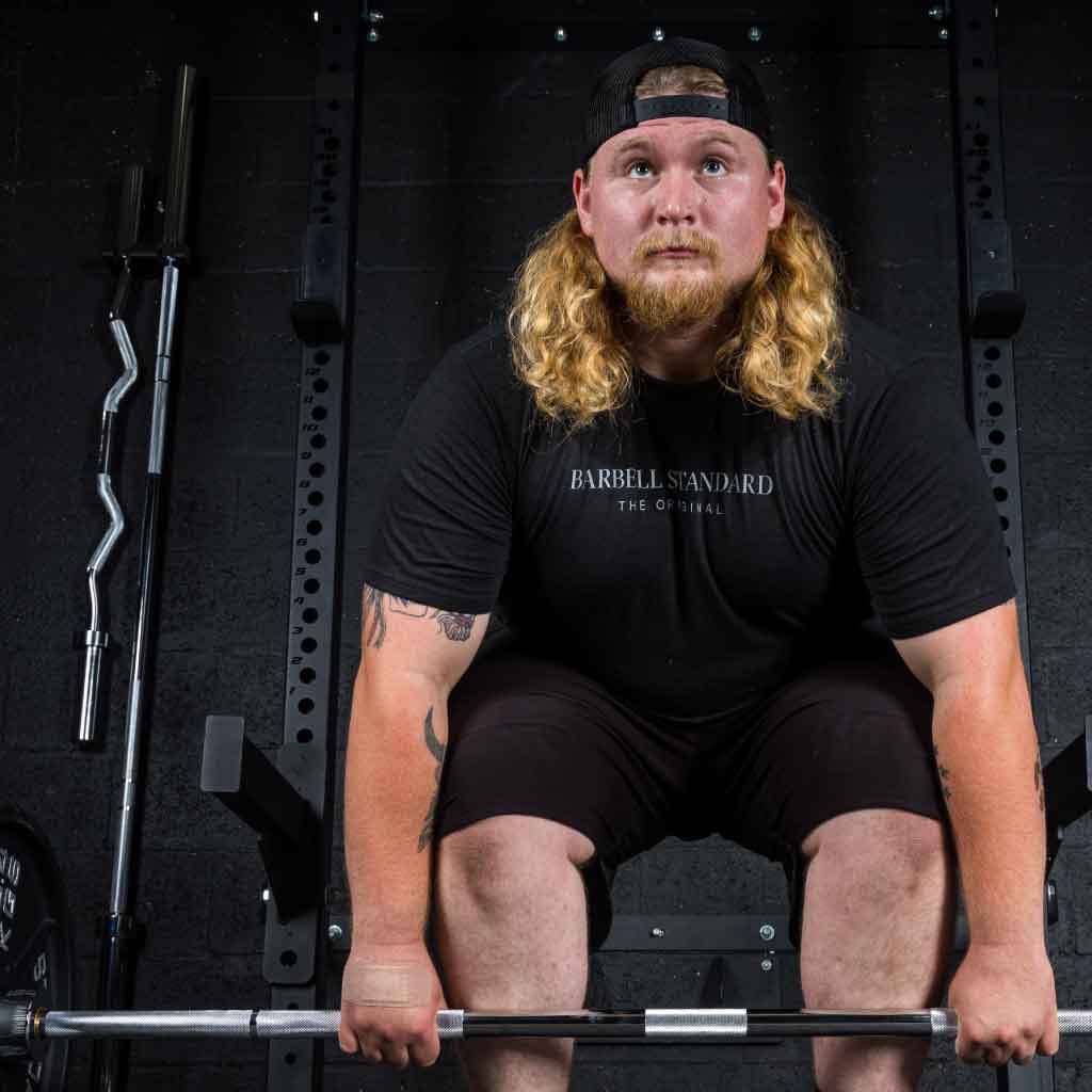 Athlete with long hair and cap preparing to deadlift Barbell Standard Olympic bar loaded with plates inside gym rack