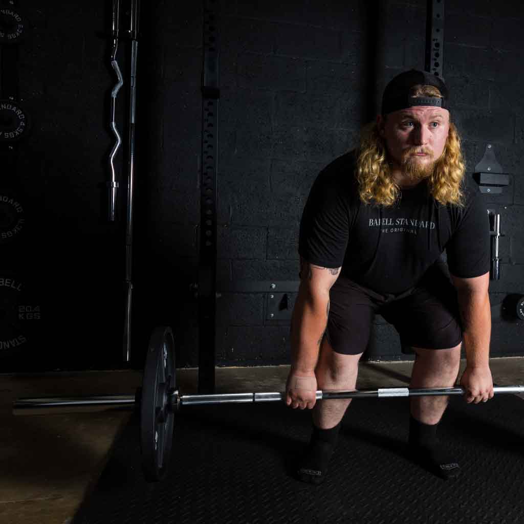 Athlete with long hair and cap preparing to deadlift Barbell Standard Olympic bar loaded with plates inside gym rack