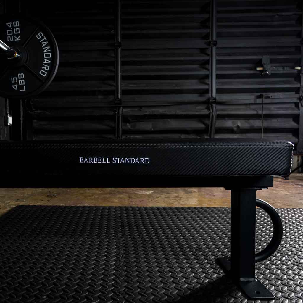 Barbell Standard competition flat bench with carbon fiber style pad and 45 lb plate loaded barbell visible on the left