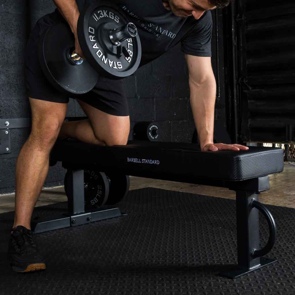 Man performing dumbbell rows on Barbell Standard competition flat bench using 25 lb iron weight plates