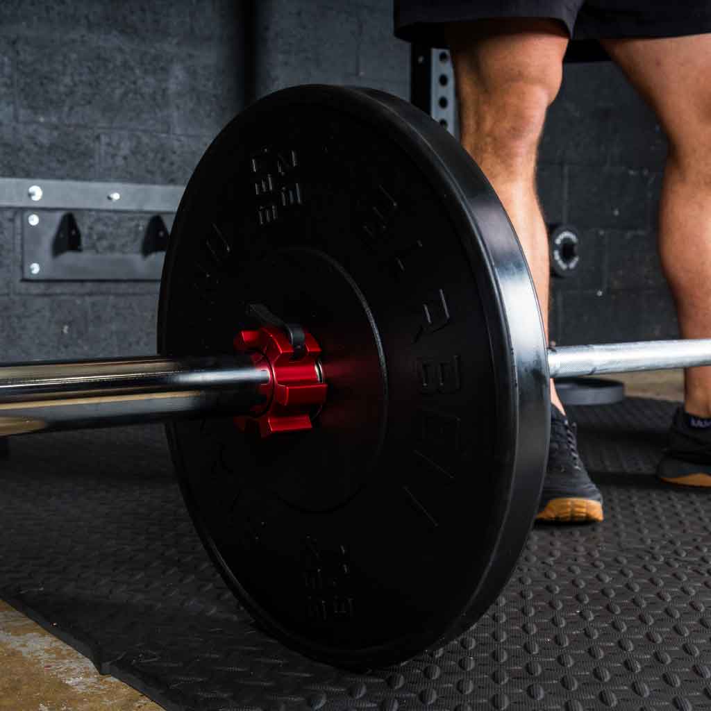 Barbell with 45 lb rubber bumper plates and red collar clamps resting on gym floor with athlete preparing to lift