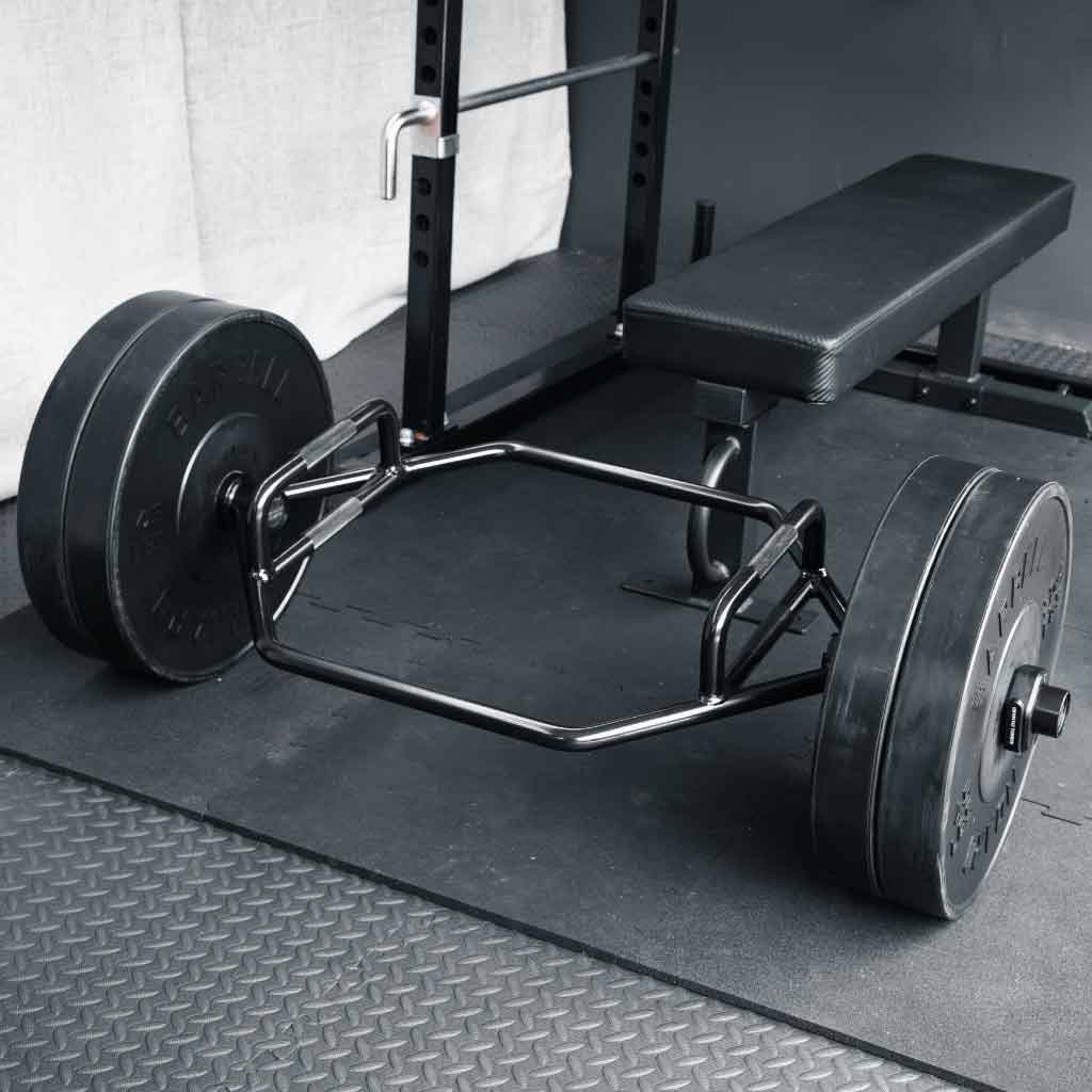 Barbell Standard Trap Bar 2.0 with weight plates on gym floor next to flat bench