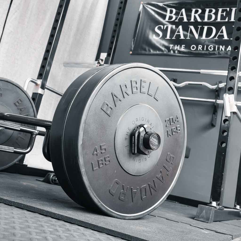 Barbell Standard trap bar loaded with 45 lb weight plates secured by magnetic collars, set up in a gym with a power rack and banner in the background