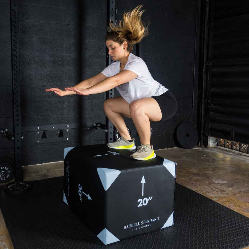 Woman performing squat jump landing on Barbell Standard 20-inch foam plyo jump box during workout in gym