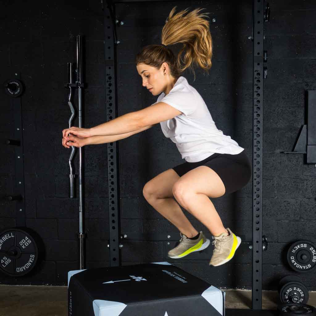 Woman mid-air performing box jump onto Barbell Standard 3-in-1 foam plyo jump box in gym setting