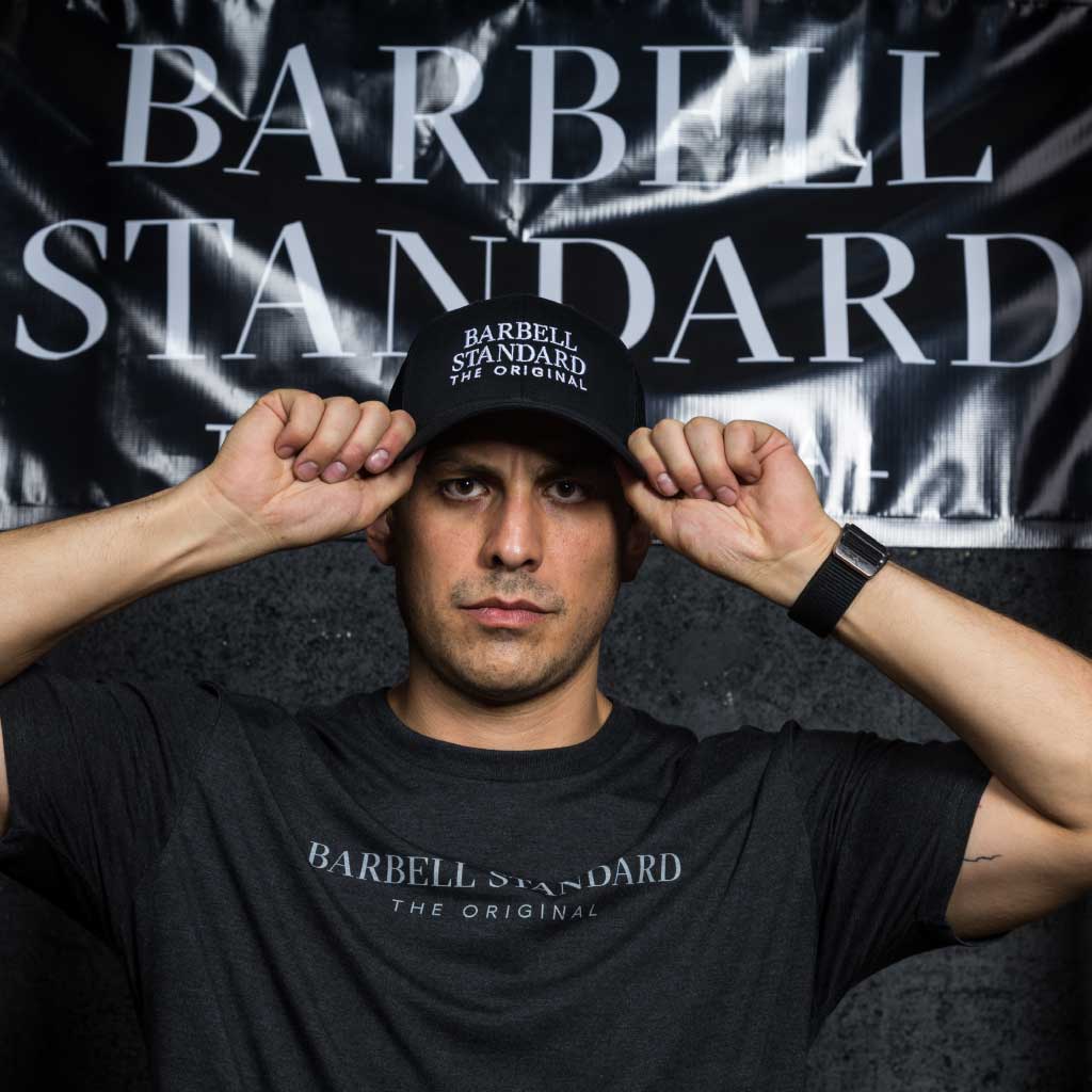 Man wearing black Barbell Standard trucker hat and t-shirt with white logo in front of Barbell Standard banner