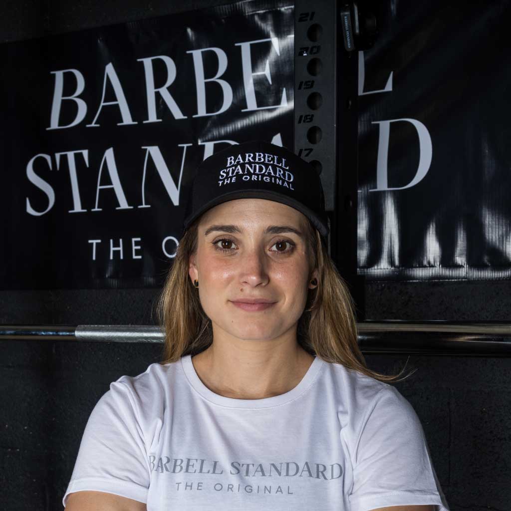 Woman wearing black Barbell Standard trucker hat and white Barbell Standard t-shirt in front of branded banner