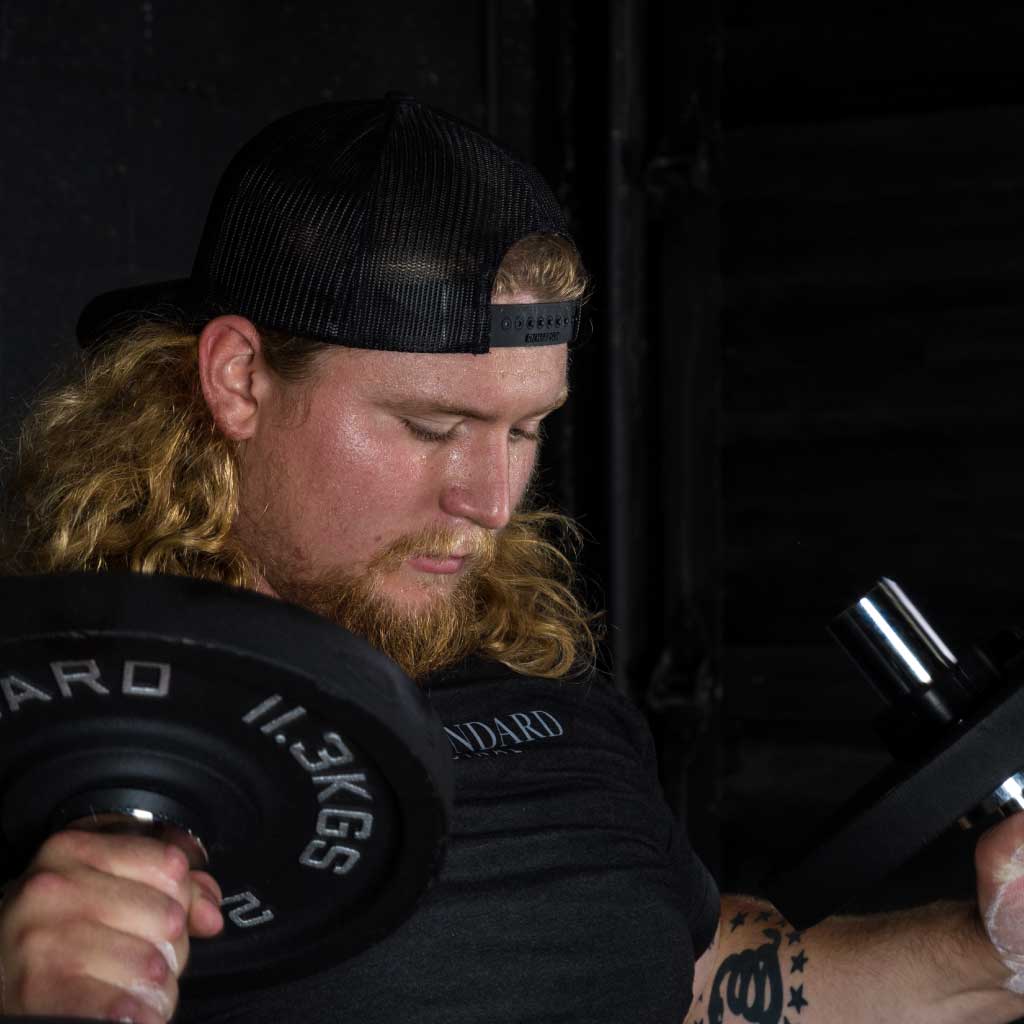 Man lifting dumbbell while wearing black Barbell Standard trucker hat and t-shirt