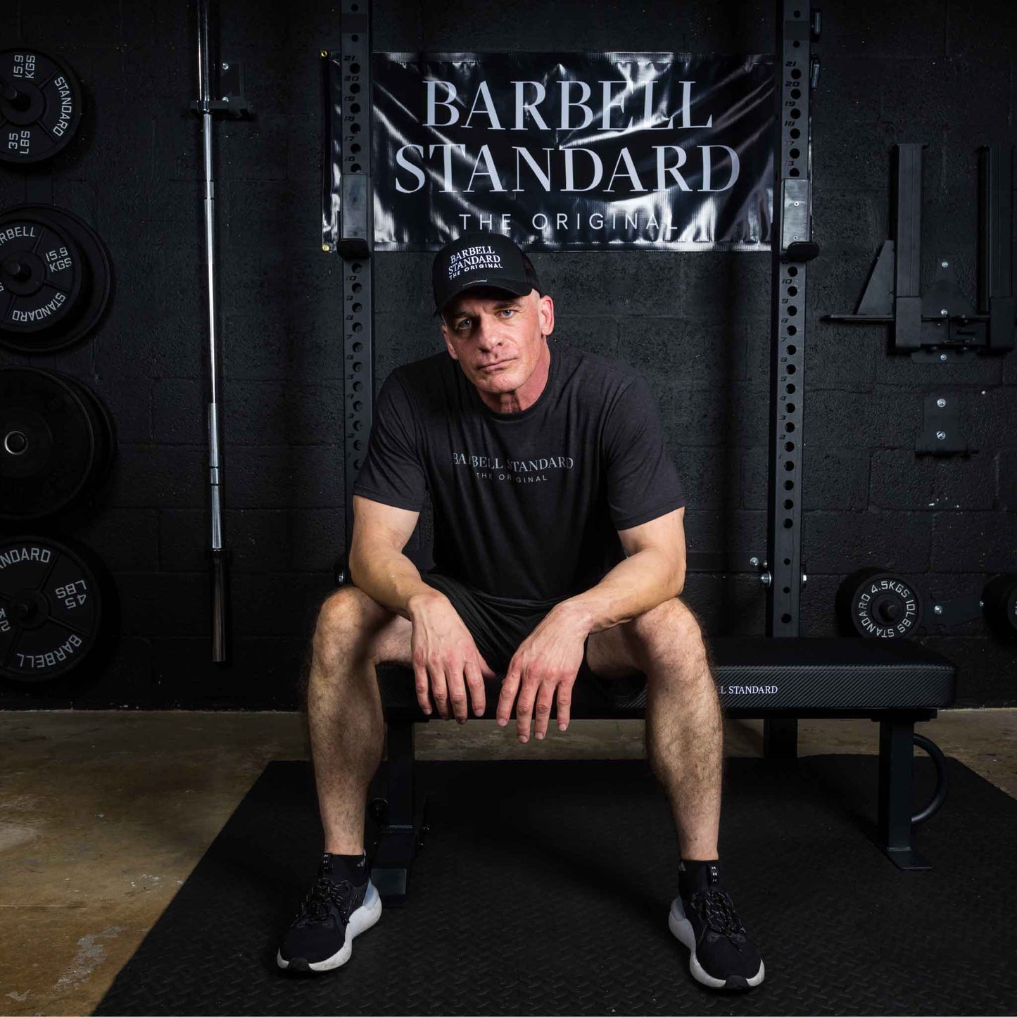 Man wearing Barbell Standard shirt and hat sitting on flat weight bench in gym with Barbell Standard banner behind