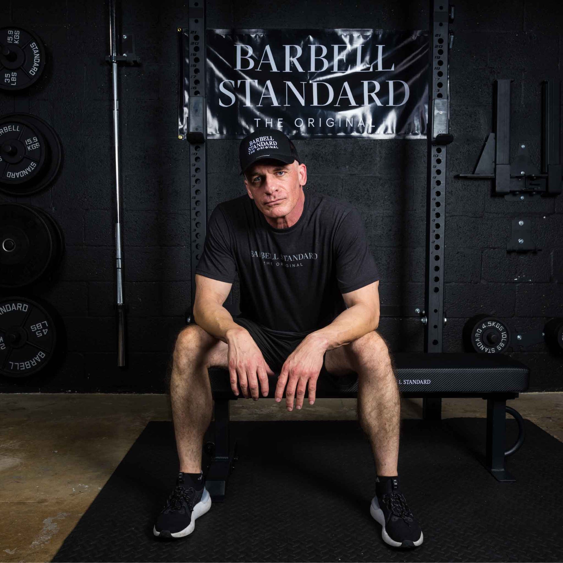 Man wearing Barbell Standard shirt and hat sitting on flat weight bench in gym with Barbell Standard banner behind
