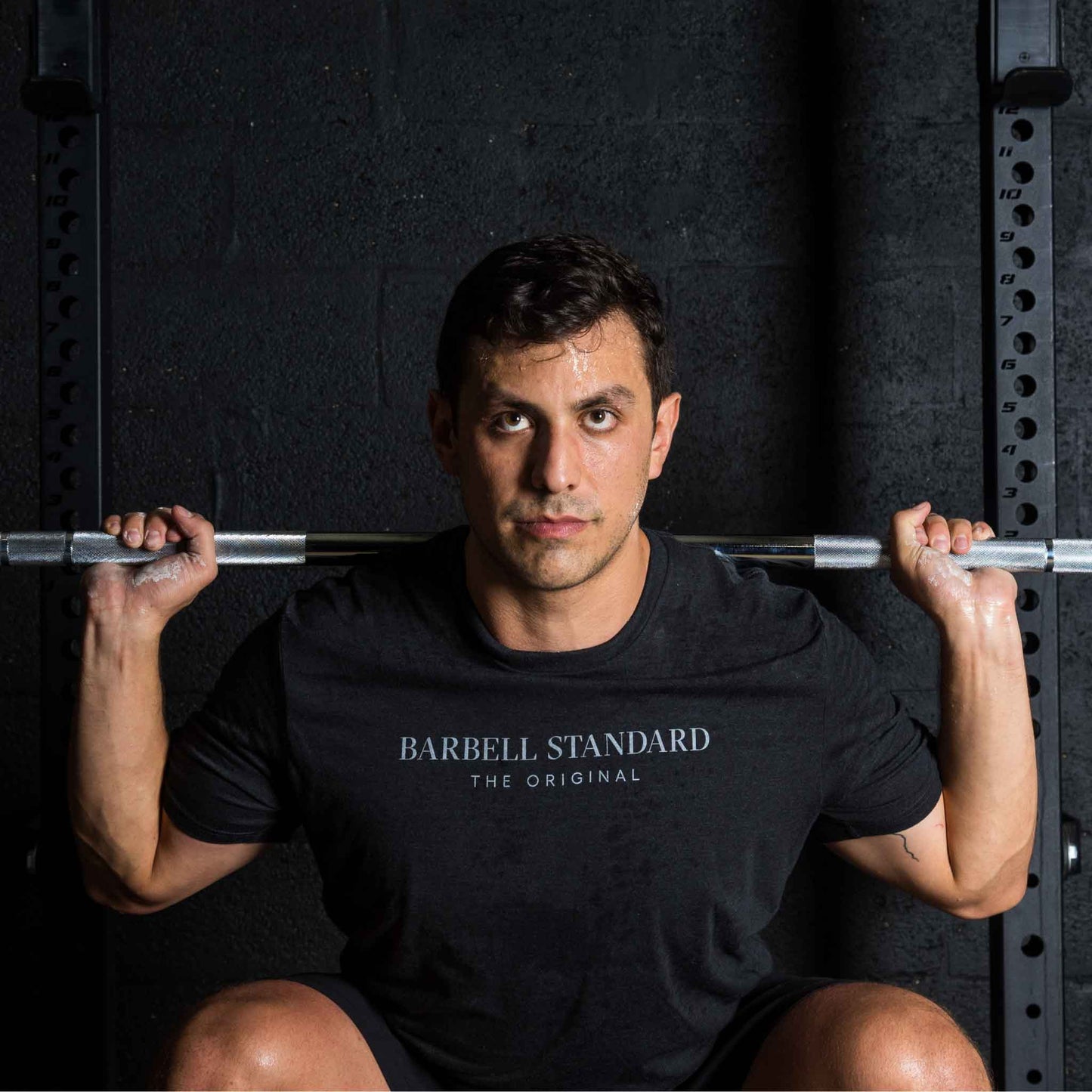 Man performing a squat with a barbell across his shoulders, wearing a black "Barbell Standard The Original" t-shirt, in a gym setting with a dark wall and squat rack