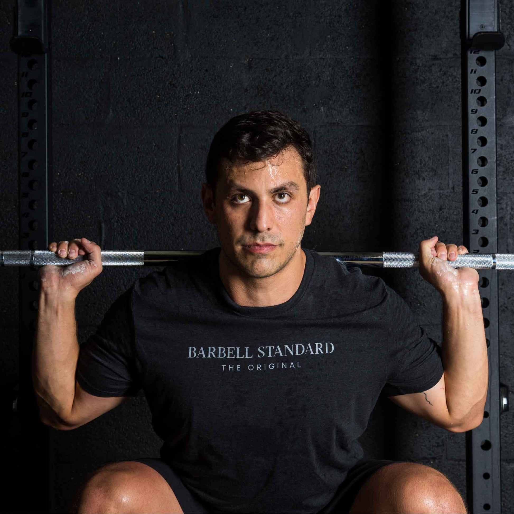 Man performing a squat with a barbell across his shoulders, wearing a black "Barbell Standard The Original" t-shirt, in a gym setting with a dark wall and squat rack