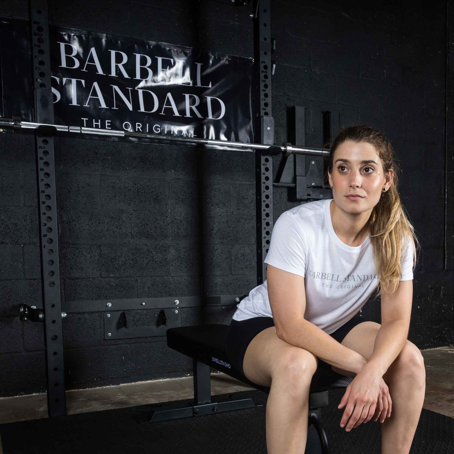 Woman in Barbell Standard shirt sitting on flat weight bench in gym with Barbell Standard banner in background