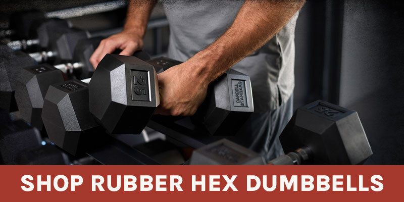 A close-up lifestyle shot of a person in a gym holding a black hexagonal dumbbell with a silver weight plate insert. Other dumbbells are visible on a rack in the background.